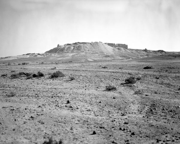 A wide, scrub-covered desert landscape with a prominent archaeological mound and stone ruins centered on the horizon.