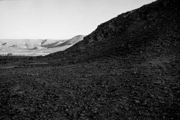 Ariel Yannay: The Mountain of God. A high-contrast view of a dark, rocky foreground slope with pale, layered mountains visible in the far distance.