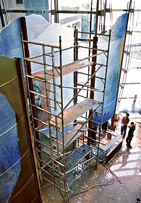Large dichroic glass sculpture like an oversize modular screen (green, yellow, blue) with a construction scaffolding and men working