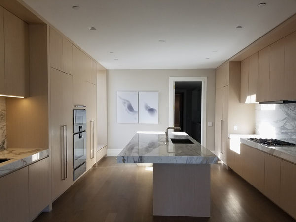 An interior kitchen with marble countertops and high-end wooden cabinets. Artwork hangs on the wall.