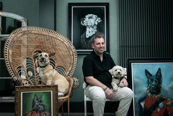 A man sitting with two dogs surrounded by dog portraits in a gallery setting.