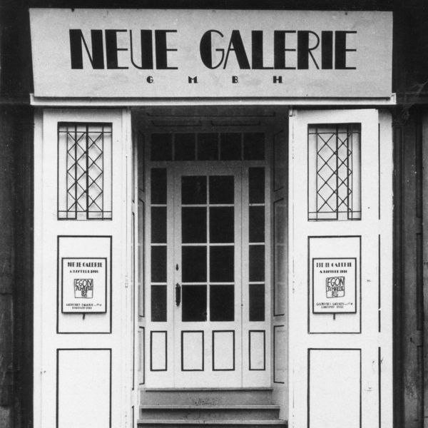 a black and white photograph of the exterior view of the entrance to the Neue Galerie in Vienna, Austria: a window paneled white door accented with black trim. posters for an Egon Schiele exhibition hang at either side