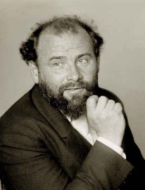A black and white photograph of Gustav Klimt: a middle-aged, balding white male in a suit, his right hand lifted to his chin as he looks off into the distance