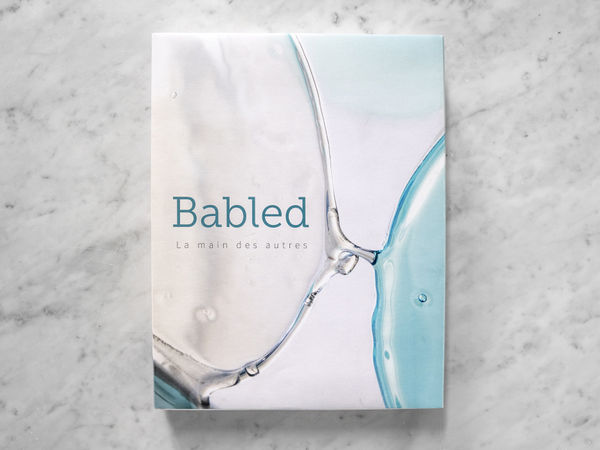 Cover of the book Babled – La main des autres: abstract detail of glass and blue liquid on a light background, evoking gesture and material. Photo by Laura Veschi.