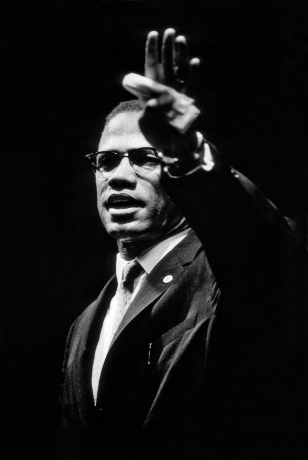 Half-length portrait of Malcom X in the stance of an orator, shown with his left hand raised, palm out, with what looks like a folded handkerchief held down with his thumb.