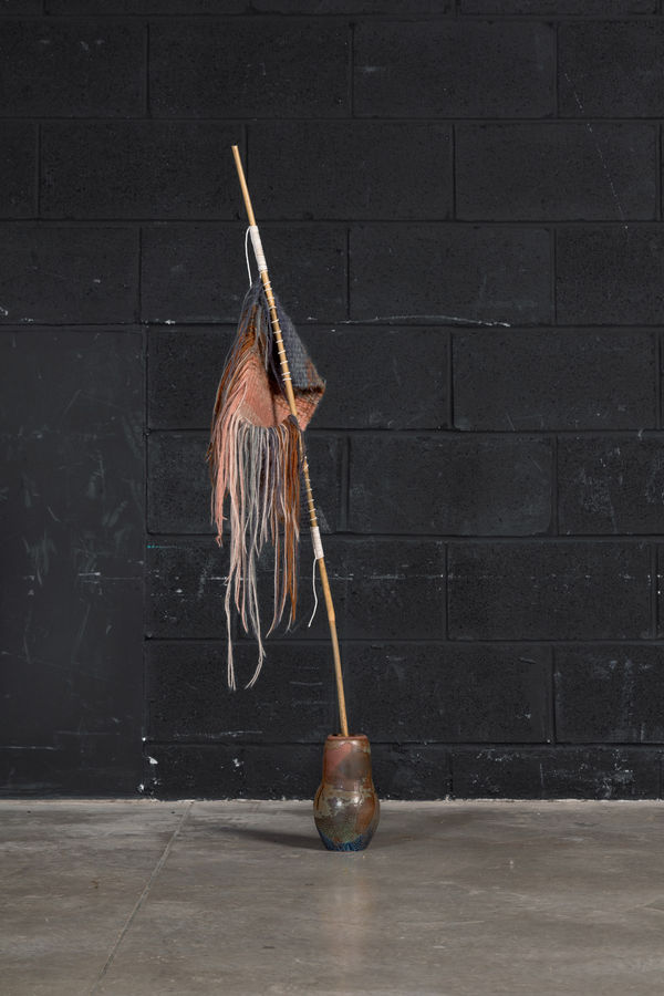 Hand formed ceramic vessel with a bamboo stick protruding from the opening. Attached to the bamboo is a hand knit swatch of rust coloured wool with many strands of wool hanging from it.