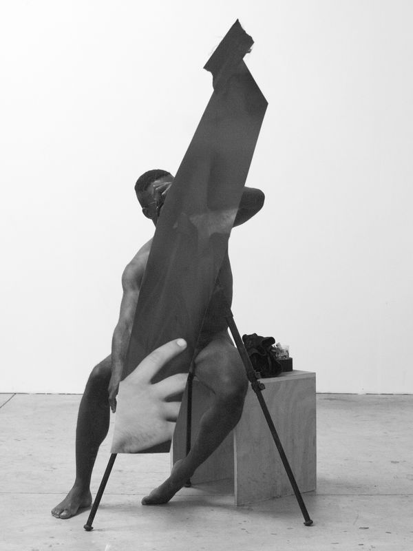 Seated Black man in a photography studio, holding a large shard-like black shape that obscures most of his nude body.
