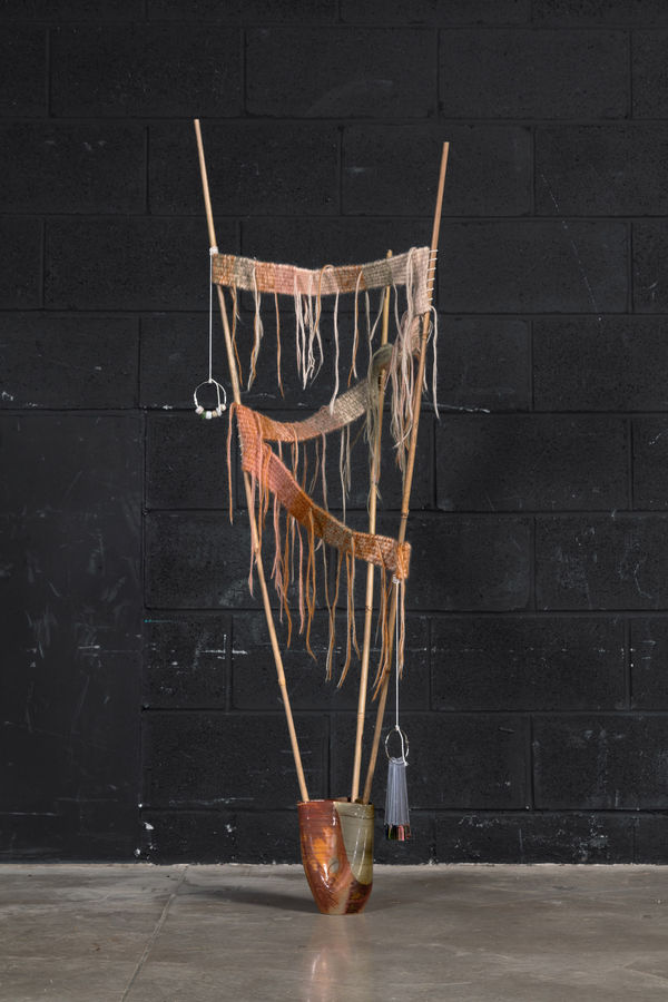 A hand made ceramic vessel with three bamboo sticks coming out of the three openings at the top. The sticks have a hand knit textile zigzagging between them. From the top of one bamboo stick hangs a key ring with plastic beads on it. From near the bottom 