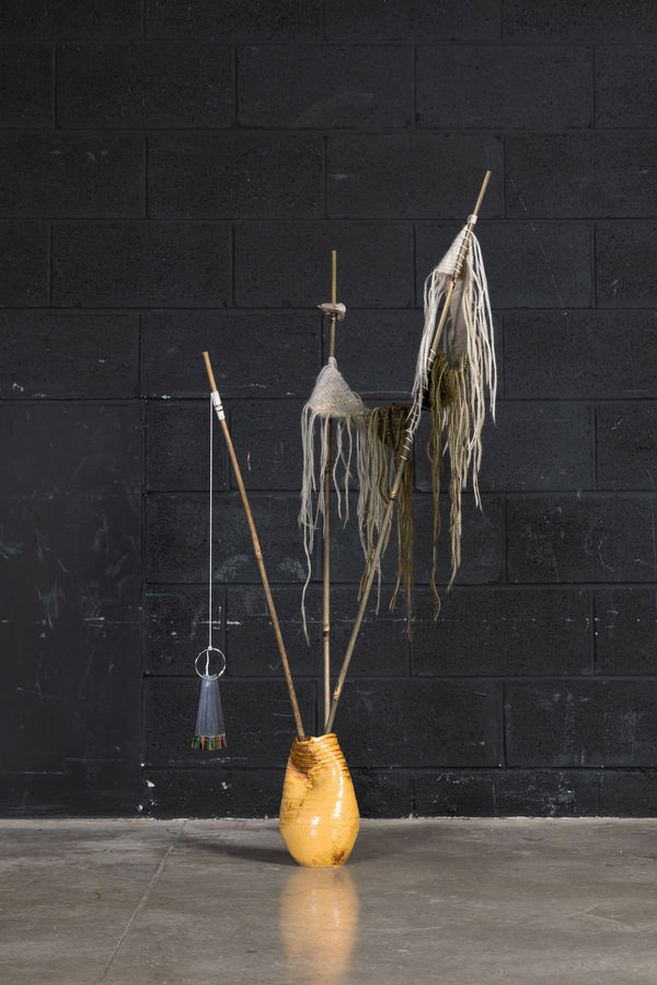 Hand built ceramic vessel with three bamboo sticks protruding from the opening. One stick has a nail polish sampler hanging from it. The other two sticks have a hand knit swatch running between them. The swatch has many loose threads hanging from it.