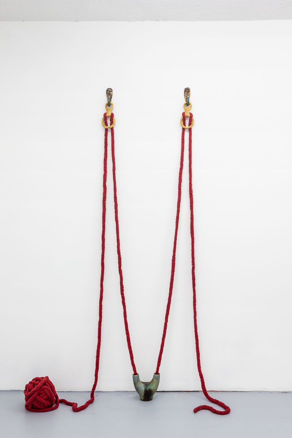 a ball of thick red yarn sits on the floor but has been pulled up over a handmade ceramic hook which hangs about 5 feet from the floor, then back down and into one opening of a hand made ceramic vessel that sits on the floor. The yarn then loops out the s