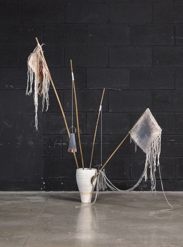 A hand made ceramic vessel with three bamboo sticks protruding from the top opening. A fourth bamboo stick is protruding from an opening on the side of the vessel. The two outer sticks have hand knit wool swatches hanging from them which contain many loos