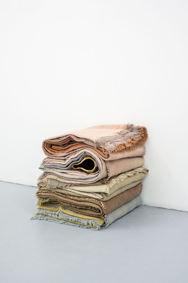 Stack of hand dyed sheets of cotton which have been folded and stacked on to of one another. two thirds of the way up the stack, a handmade ceramic vessel has been placed between the layers so that the edge and opening are visible on one side of the stack