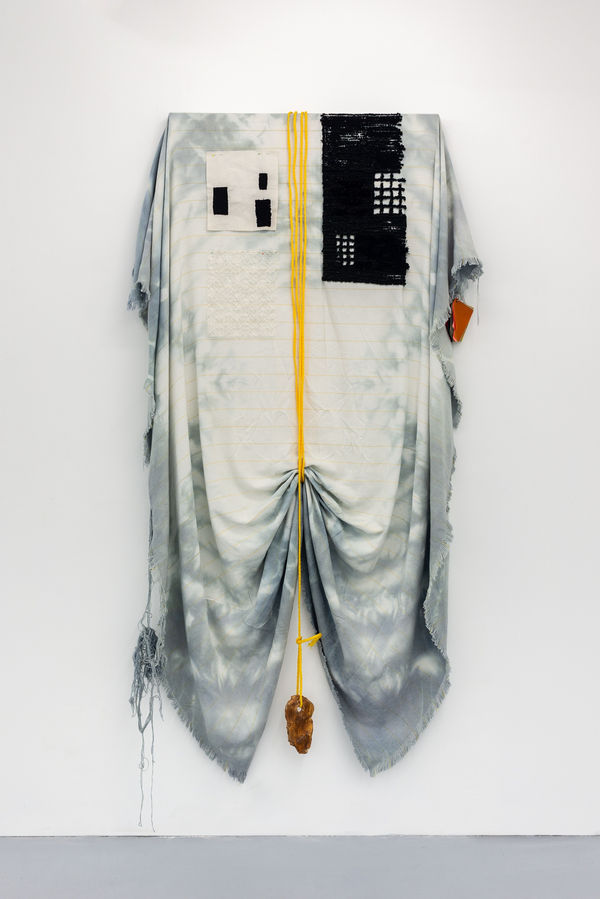 A large sheet of hand died monks cloth hangs from a wooden painting stretcher. The cloth hangs down below the bottom of the stretcher and over both sides. The cloth is sinched at the mid point of the bottom by a nylon rope that wraps around the frame, ove