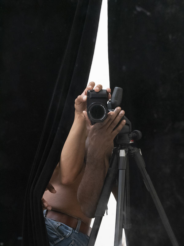 Black drapery is parted to reveal a camera on a tripod aimed at us, the viewer. The camera is being held by a black arm and a white arm that emerge from the drapery, the rest of the two bodies concealed behind the black drapery.