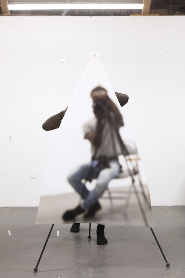 View of a photographer seated in a studio aiming a camera out at us, mostly obscured by a cut-out of a photograph of another photographer also aiming at us.