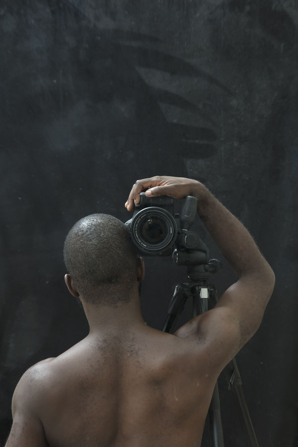 View of a shirtless Black photographer seen from behind, his right arm holding a camera at his shoulder aimed at us, the viewer.