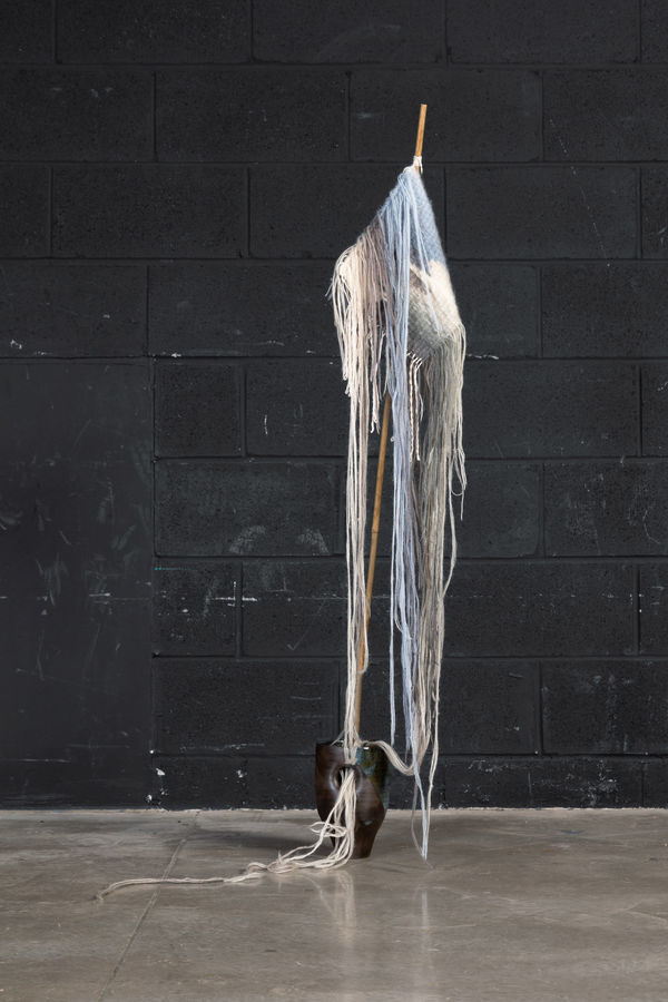 A hand build ceramic vessel with a bamboo stick protruding from the top opening. Attached to the stick is a hand knit wool swatch with many long loose threads hanging from it. Some of these long threads hangs down and flow back through the top opening and
