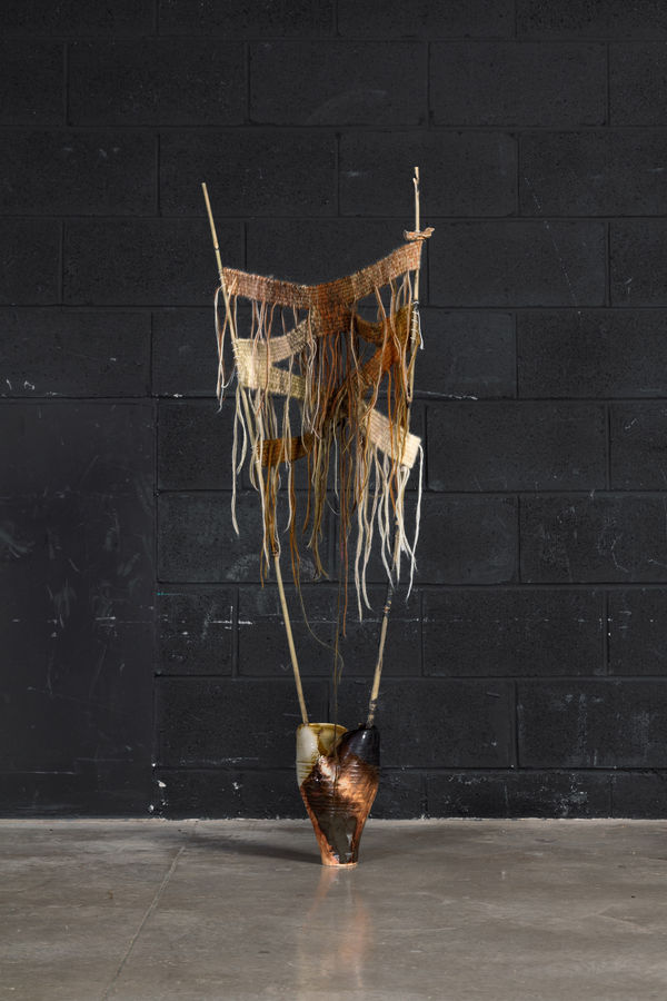 A hand made ceramic vessel with two bamboo sticks coming out of the opening at the top. The sticks have a hand knit textile zigzagging between them