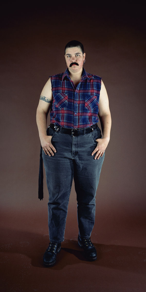 Full-length self-portrait of Catherine Opie, wearing jeans, a sleeveless flannel shirt, and a fake black mustache.
