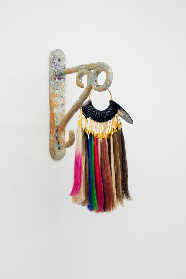 An ornate ceramic hook hangs from the wall. Attached to the end of the hook is a multicoloured sampler of different shades of synthetic hair.