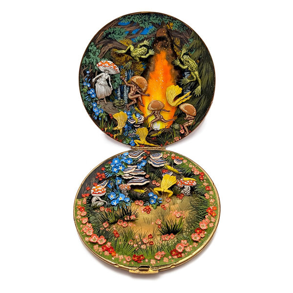 Image of an open compact filled with a scene of cutout mushroom head people dancing in a forest around a large fire underneath the trees with lots of flowers and two large frogs 