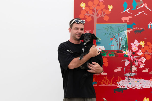 Artist Francisco Díaz Scotto holds a small black dog, Meera, in front of his large red painting on a white wall.