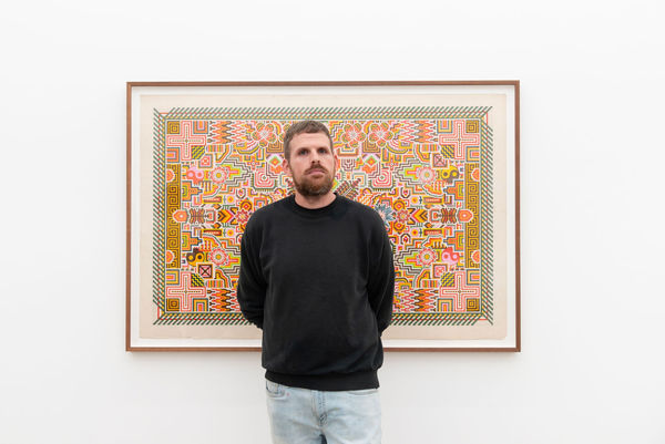 Artist Matthew Craven, a white man with short brown hair and a black sweater, stands in front of his intricately drawn pattern, DREAM/WEAVR, a framed work on found paper