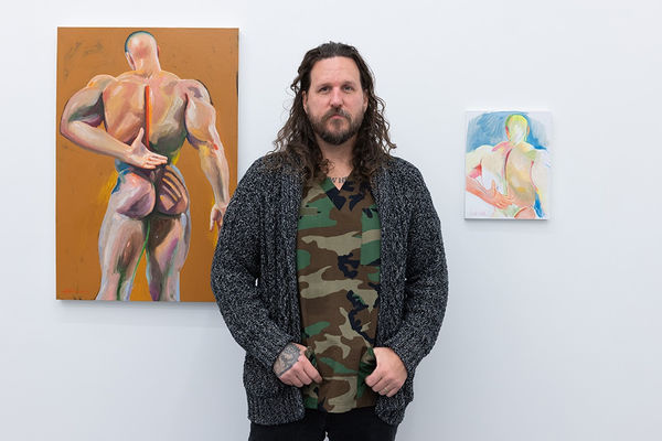 Erik Jones, a white man with long hair wearing a army camo print shirt, stands between his two artworks, one painting and one study drawing, of the backside of a muscular man. He stands in a white cube art gallery with white walls.