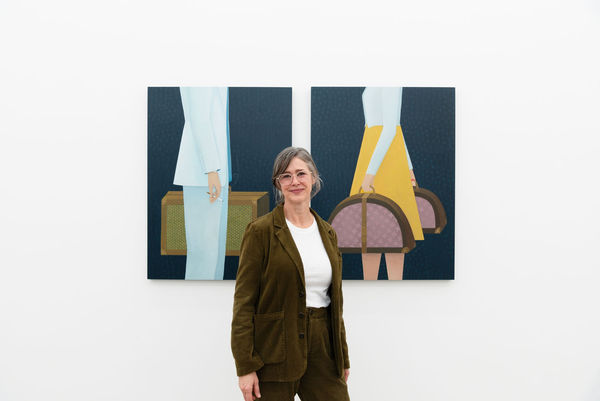 Artist Angela Burson, a white woman with grey hair, stands in front of her two large paintings hung on white walls in an art gallery. The paintings depicted headless figures in motion holding suitcases.