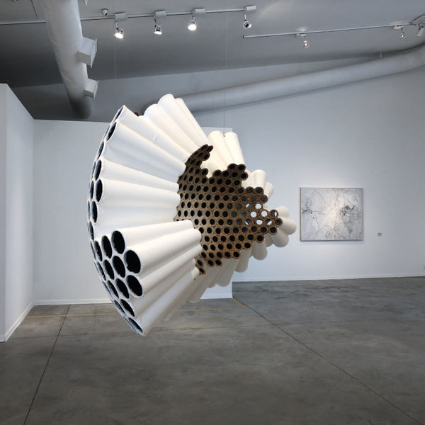 Installation view of a white, black, and gold honeycomb-like sculpture hanging from the ceiling with a white-and-black rectangular work hanging on the wall in the background.