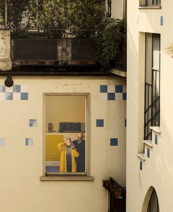 Gail Albert Halaban Out My Window Italy, older man and woman in yellow jacket viewed through apartment window in light yellow building
