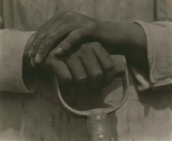 a worker's worn, dusty hands rest upon the handle of a tool, against a backdrop of soiled work clothes, by Tina Modotti