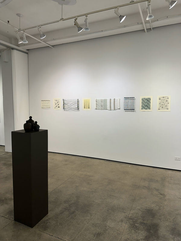 photo of an installation with three small urns on a pedestal. behind them on the wall are a series of small works.