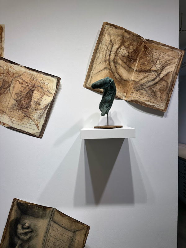 Photo of an installation, with one sculpture sitting on a platform emerging from the wall. Behind it, are three two page spreads with images drawn over the text, placed irregularly on the wall.