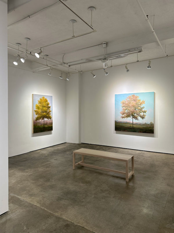 Photo of an installation with two paintings visible. They are of trees, framed symmetrically in the center of the canvases. There is a bench in the center of the room.