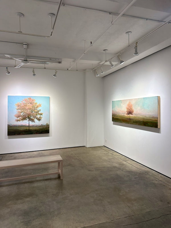 Photo of an installation with two paintings visible. They are of trees, framed symmetrically in the center of the canvases. There is a bench in the center of the room.