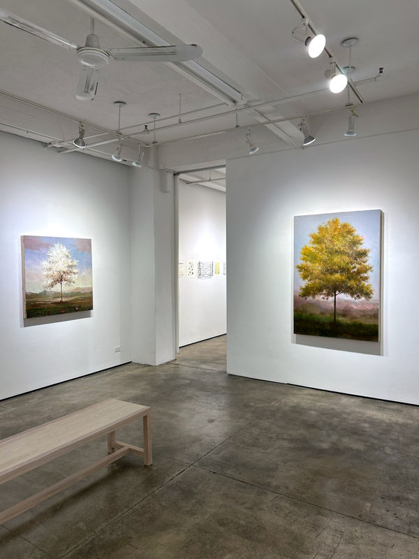 Photo of an installation with two paintings visible. They are of trees, framed symmetrically in the center of the canvases. Between the two paintings is a doorway into another room.
