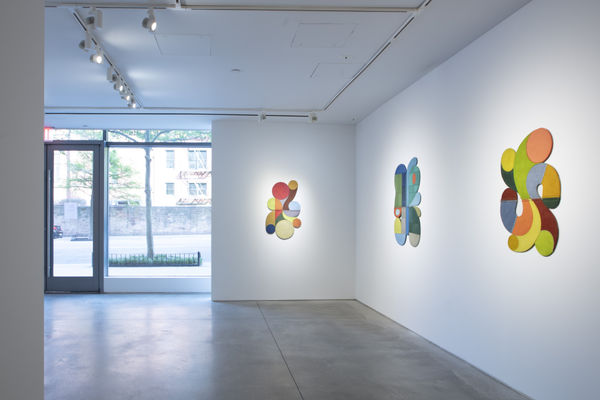 Photo of an exhibition, with three works visible on the walls. They are flat shallow sculptures, with circular motifs in dark and light shades of colors. The exhibition wall ends, and there is a large window and glass door way which go from the floor to t