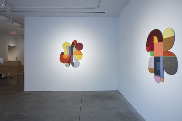 Photo of an exhibition, with two works visible on the walls. They are flat shallow sculptures, with circular motifs in dark and light shades of colors. The white wall ends before the left border of the picture, and there is a desk visible behind the wall.