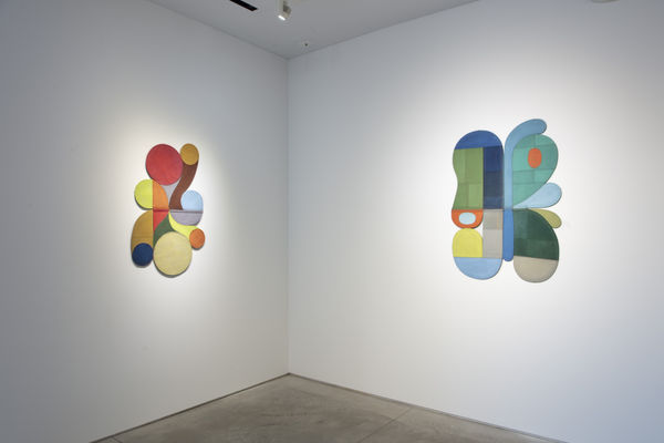 Photo of an exhibition, with two works visible on the walls. They are flat shallow sculptures, with circular motifs in dark and light shades of colors.