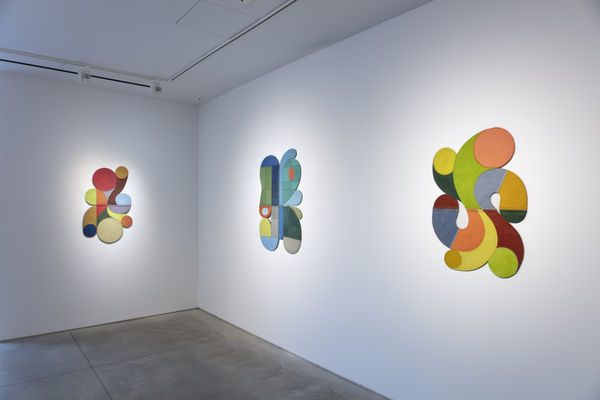 Photo of an exhibition, with three works visible on the walls. They are flat shallow sculptures, with circular motifs in dark and light shades of colors.