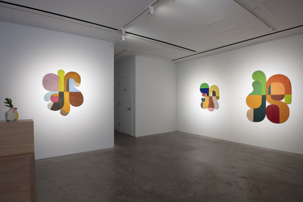 Photo of an exhibition, with three works visible on the walls. They are flat shallow sculptures, with circular motifs in dark and light shades of colors. There is desk porting into the  frame of the photo on the left side.