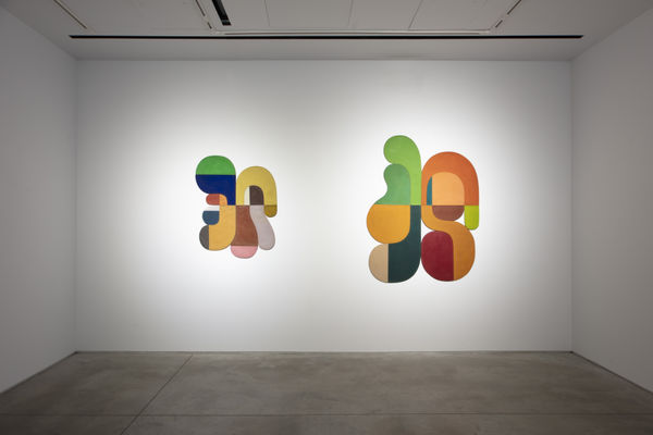 Photo of an exhibition, with two works visible on the walls. They are flat shallow sculptures, with circular motifs in dark and light shades of colors.