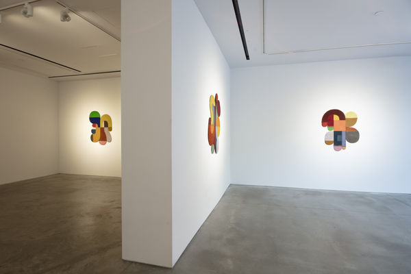Photo of an exhibition, with three works visible on the walls. They are flat shallow sculptures, with circular motifs in dark and light shades of colors. One work is on a wall perpendicular to the other, creating a barrier between the other two works.