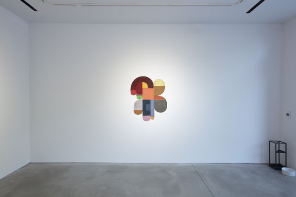 Photo of an exhibition, with one works visible on the wall. It is a flat shallow sculpture, with circular motifs in dark and light shades of colors.