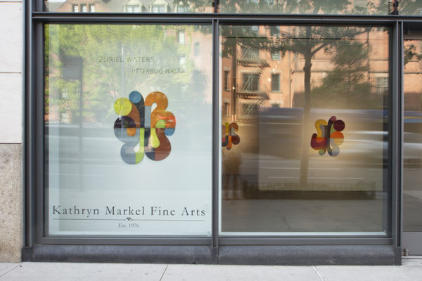 Photo of an exhibition from Kathryn Markel Fine Arts, from outside. There are two large window panels which go from floor to ceiling, with the left one being covered by a white wall behind it and the right looking into the show. There is one work visible 