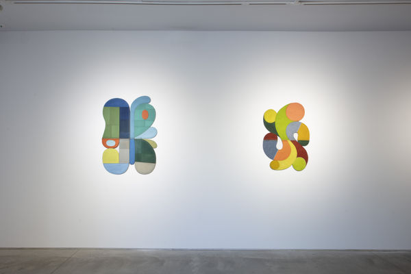 Photo of an exhibition, with two works visible on the walls. They are flat shallow sculptures, with circular motifs in dark and light shades of colors.