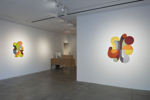 Photo of an exhibition, with two works visible on the walls. They are flat shallow sculptures, with circular motifs in dark and light shades of colors. The walls are perpendicular, But there is a space between them, in which a brown desk sits.