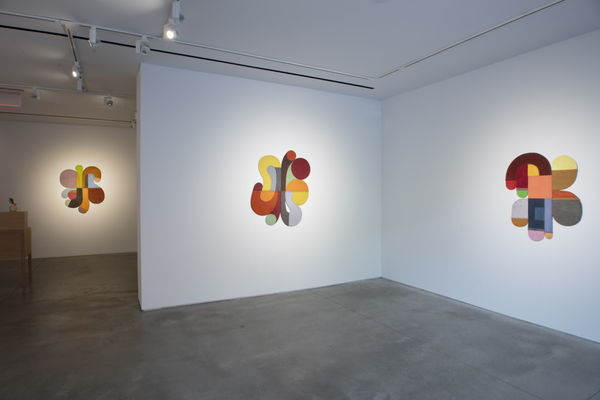 Photo of an exhibition, with three works visible on the walls. They are flat shallow sculptures, with circular motifs in dark and light shades of colors. There is a desk visible in the left edge of the picture.