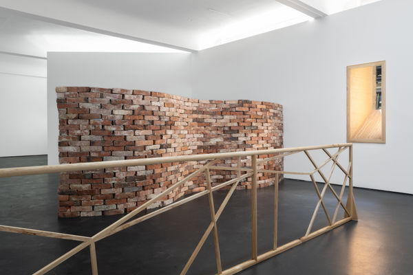 Installation view five | rindon johnson | Photo: Marjorie Brunet Plaza. Crinkle crankle brick wall, a wooden railing structure with vertical and horizontal wooden balance beams and a window with an angular wooden frame.